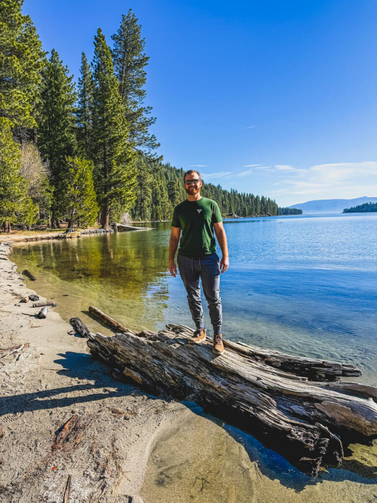 man standing on log in front of Emerald Bay in Lake Tahoe after hiking down from the top of Eagle Falls