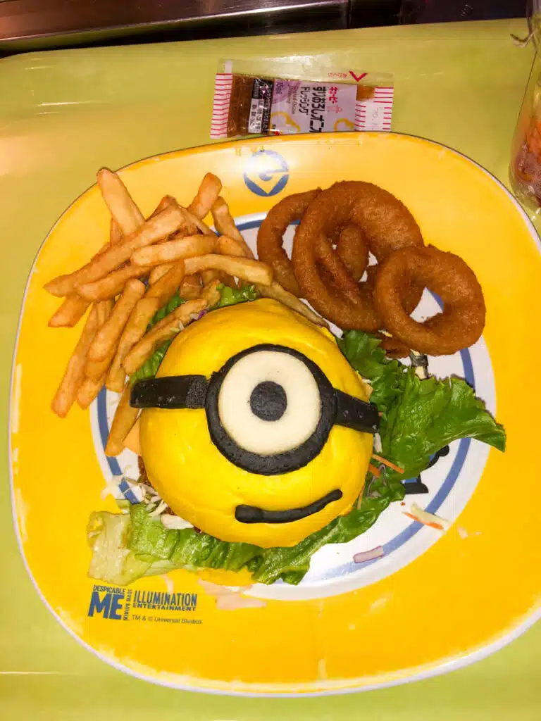 Minion themed burger meal at Universal Studios Japan in Osaka featuring a bright yellow bun designed like a Minion face with one eye served with fries onion rings and lettuce on a plate branded "Despicable Me Illumination".