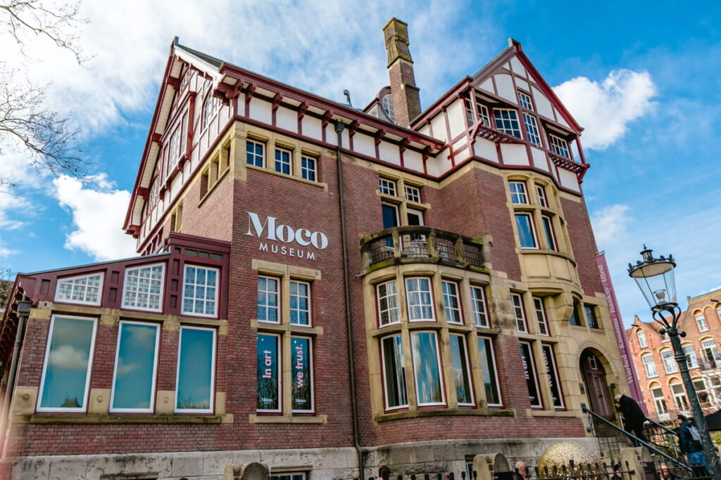 Exterior of the Moco Museum in Amsterdam with its historic brick building and large white "Moco Museum" sign on the facade under a blue sky. This museum is known for modern and contemporary art in the Museumplein area.