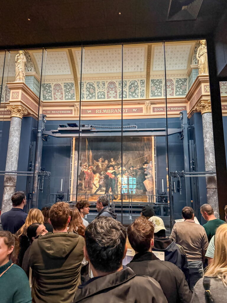 Visitors gathered inside the Rijksmuseum viewing Rembrandt’s famous painting "The Night Watch" displayed behind glass in an ornate gallery. The large artwork is framed by decorative columns and a sign above reading "Rembrandt".