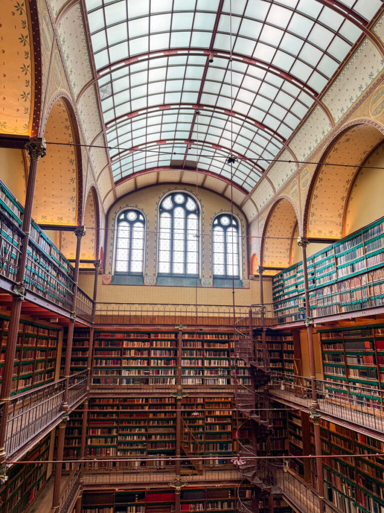 The Rijksmuseum library with multiple levels of tall bookshelves filled with books and a central spiral staircase leading upward. A glass ceiling lets in natural light, highlighting the grand historic reading room.