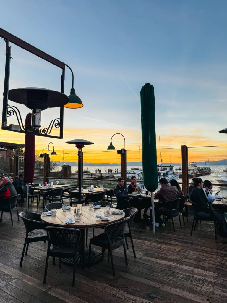 Outdoor dining patio at Riva Grill along Lake Tahoe at sunset with glowing heaters, wooden tables, and guests seated facing the water and docked boats in the background.