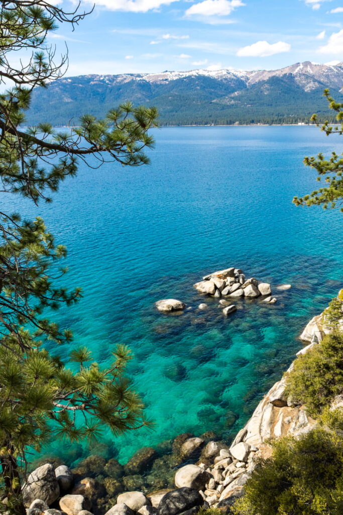 Turquoise water and large granite boulders along the rocky east shore of Lake Tahoe framed by pine branches with snow capped mountains in the distance.