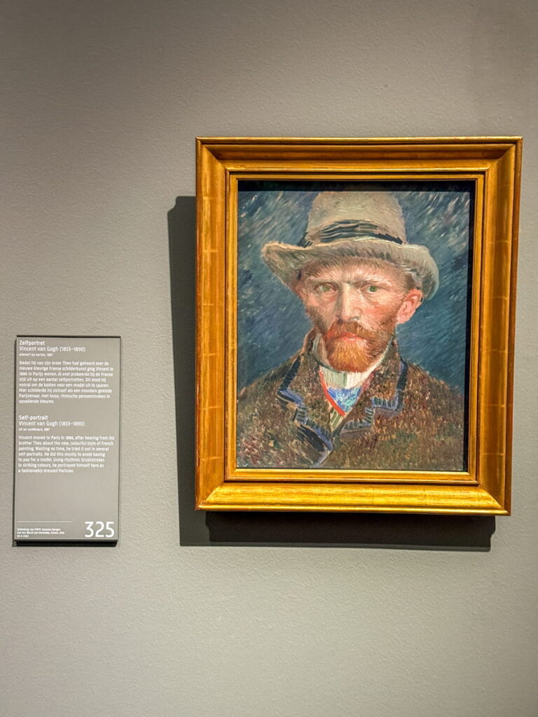 Vincent van Gogh self portrait painting displayed in a gold frame on a museum wall with an information plaque beside it reading "Self-portrait Vincent van Gogh (1853–1890)" and gallery number "325". The artwork shows the artist wearing a hat with expressive brushstrokes.