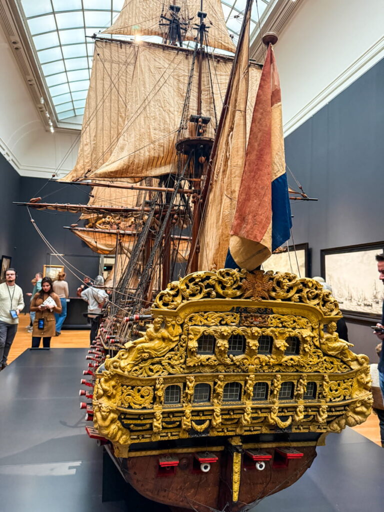 Detailed model of an ornate Dutch ship displayed inside the Rijksmuseum with intricate gold carvings and sails raised. Visitors stand nearby observing the historic maritime exhibit in the gallery.
