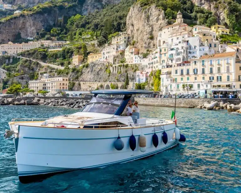 A white tour boat floats in bright blue water in front of Amalfi with pastel buildings and steep rocky cliffs rising behind it. This Amalfi Coast boat tour scene shows the town from the water with an Italian flag at the back of the boat.