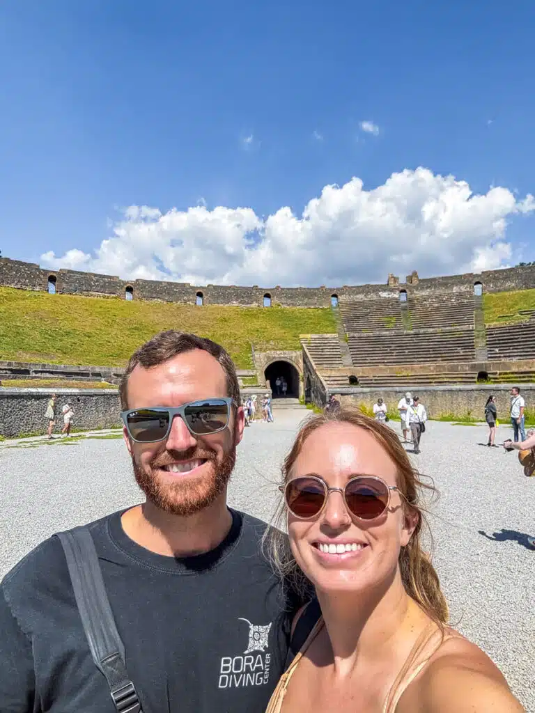 young couple smiling inside the large amphitheater of the Pompeii ruins with a blue sky and clouds in the background