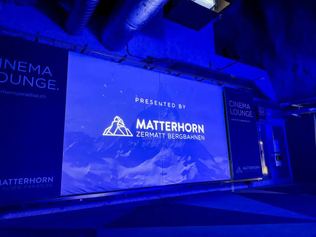 theater room lit up by a blue screen that reads "presented by Matterhorn Zermatt Bergbahnen" and "Cinema Lounge" on the sides. Taken inside the Matterhorn Glacier Paradise