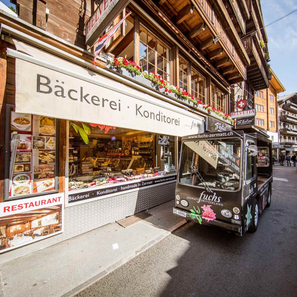 Fuchs bakery storefront in Zermatt with a Bäckerei Konditorei awning, pastry display window and a black electric Fuchs delivery cart parked outside