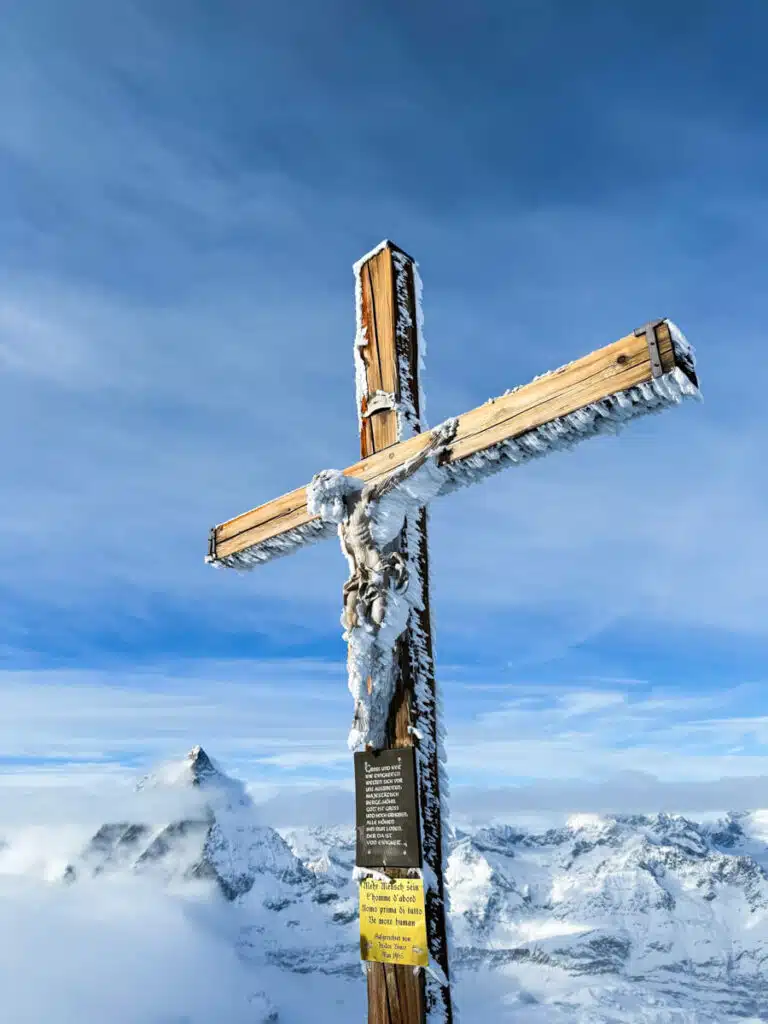 wooden cross with Christ at the top of the Matterhorn Glacier Paradise panoramic platform. The wood sculpture is covered in ice crystals and the snowy Matterhorn looms in the background