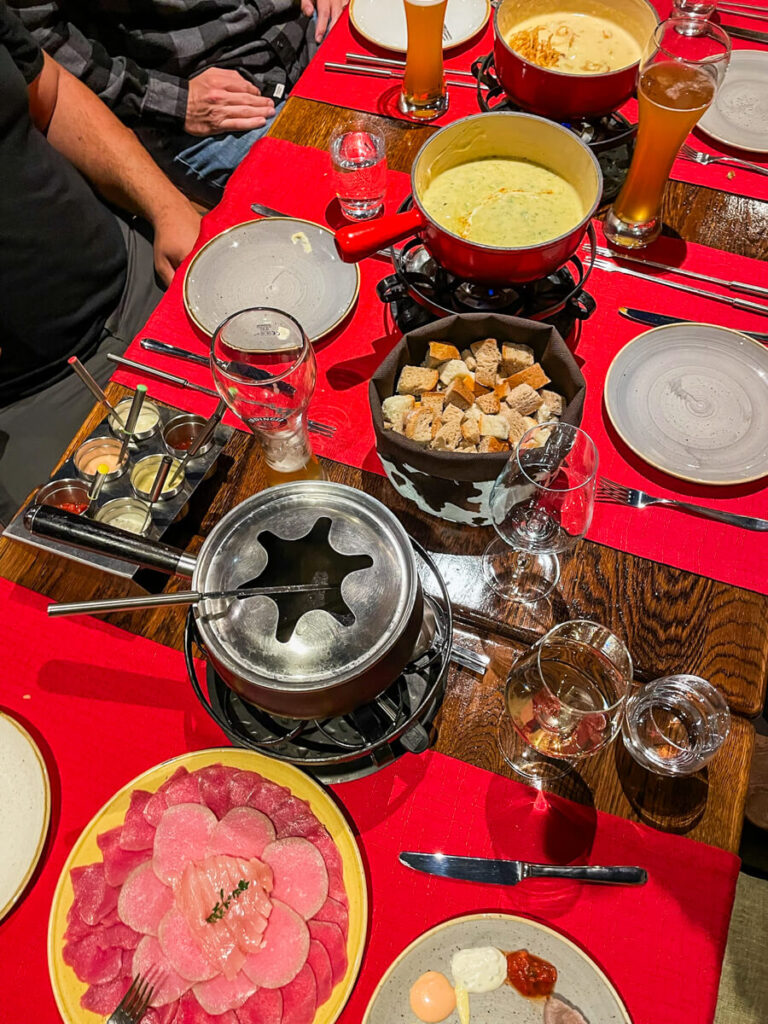 Table at Swiss Chalet restaurant in Zermatt with red pots of cheese fondue, a meat fondue pot, bread cubes, sliced raw meat and beers