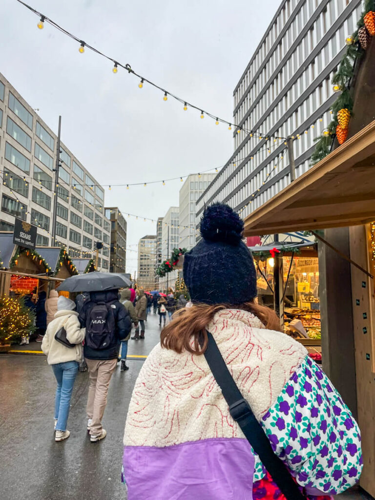 Shoppers browsing wooden Christmas market stalls at night under bare trees hung with glowing star lights