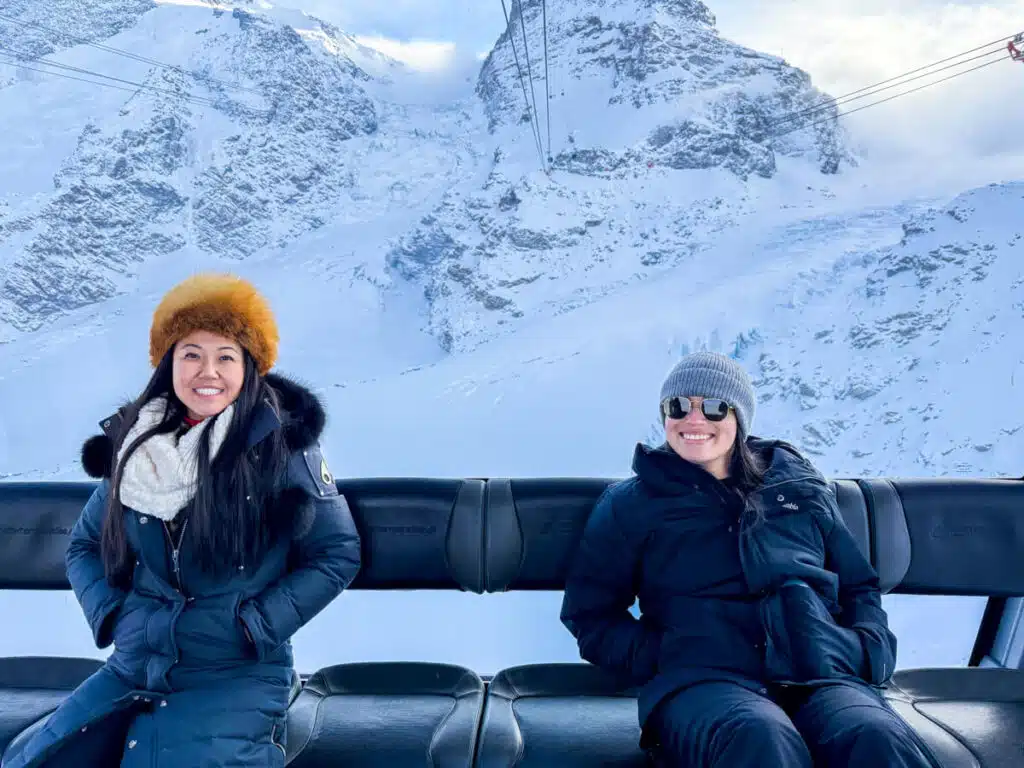 two girls in black jackets and hats sitting inside the Matterhorn Glacier Paradise gondola on the final leg up to the summit, with snowy mountains in the background