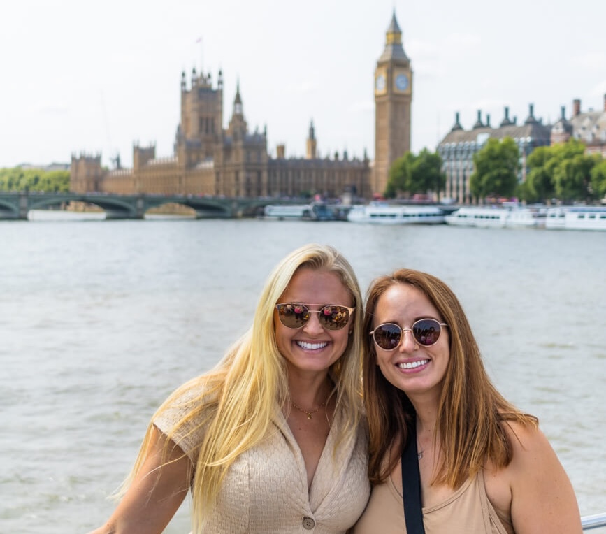 two girls in sunglasses smiling  at the camera with Big Ben and Elizabeth Tower in the background. Taken on a river cruise on the Thames in London