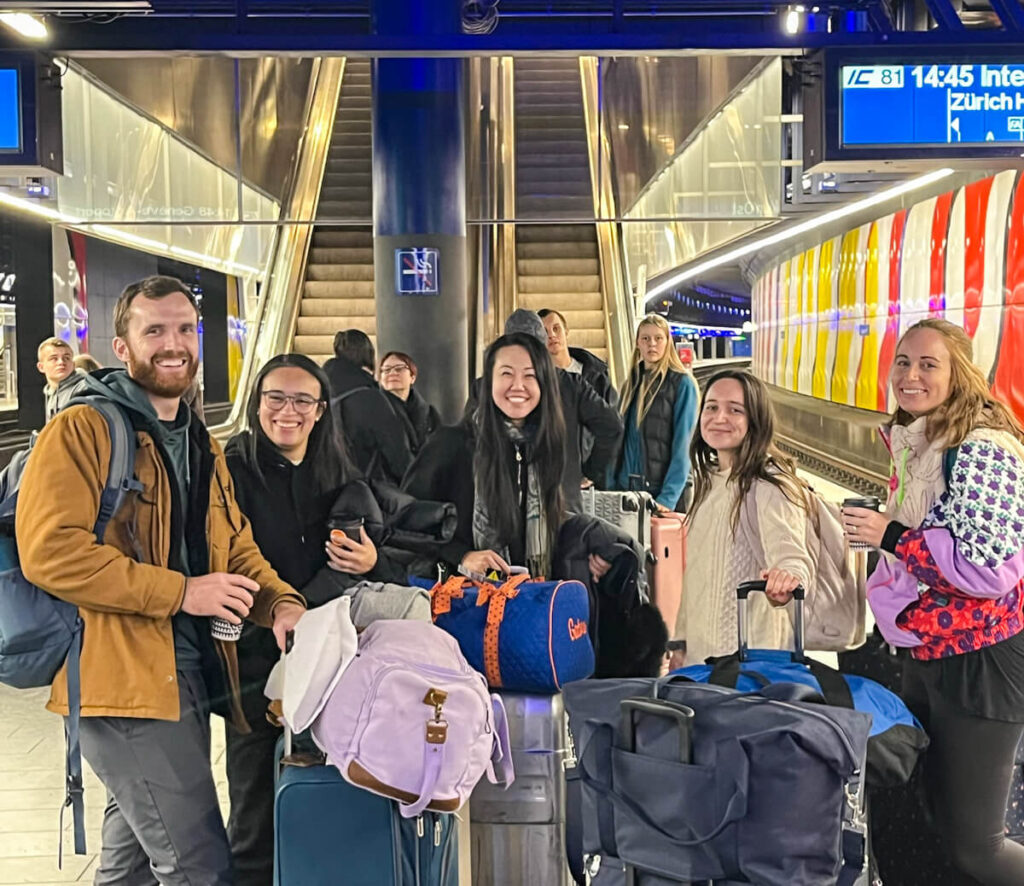 Smiling travel group posing with luggage on a Swiss train platform in Zurich, escalators and departure board behind them