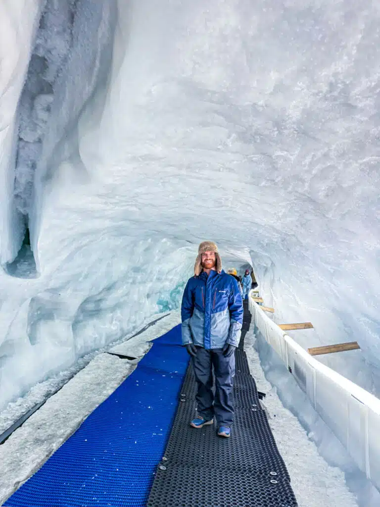 man in blue jacket and black snow pants standing inside of an ice cave on black plastic walkway next to blue carpet. Taken inside the Glacier Palace inside the Matterhorn Glacier Paradise in Zermatt, Switzerland.
