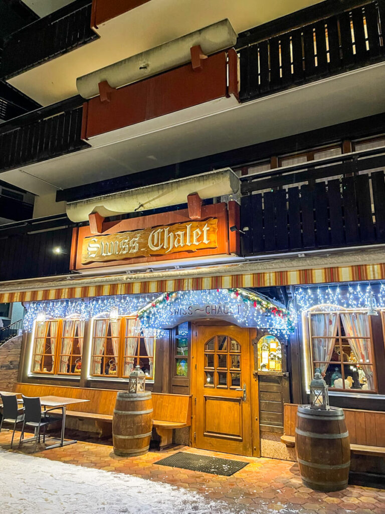 Swiss Chalet restaurant in Zermatt at night, with a carved wooden sign, wooden door and windows trimmed in twinkling Christmas lights