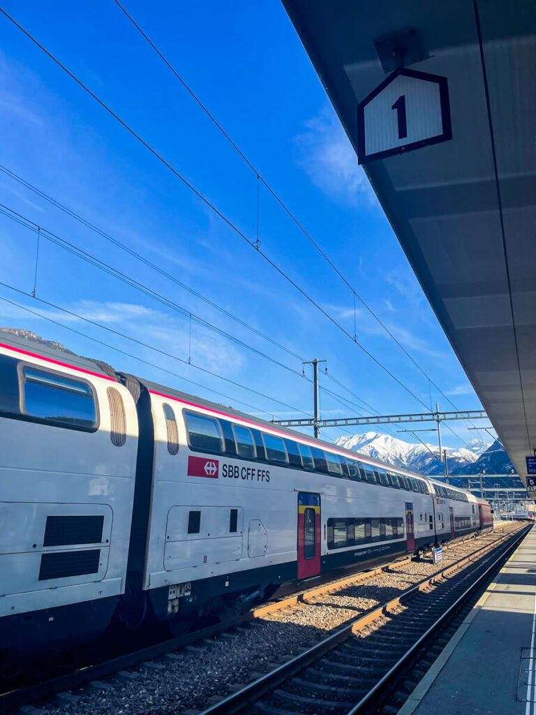Red and white SBB double-decker train at platform 1 under a blue sky, with snow-capped mountains behind