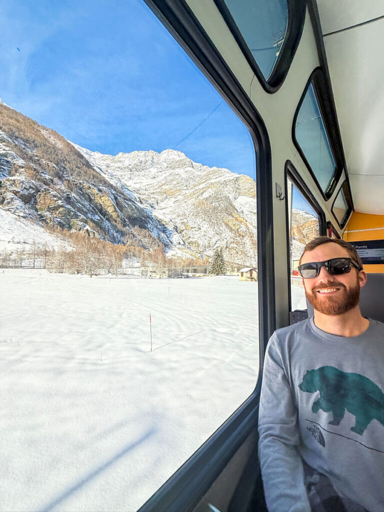 Man in sunglasses smiling beside a large train window showing snowy fields and rugged Alpine peaks