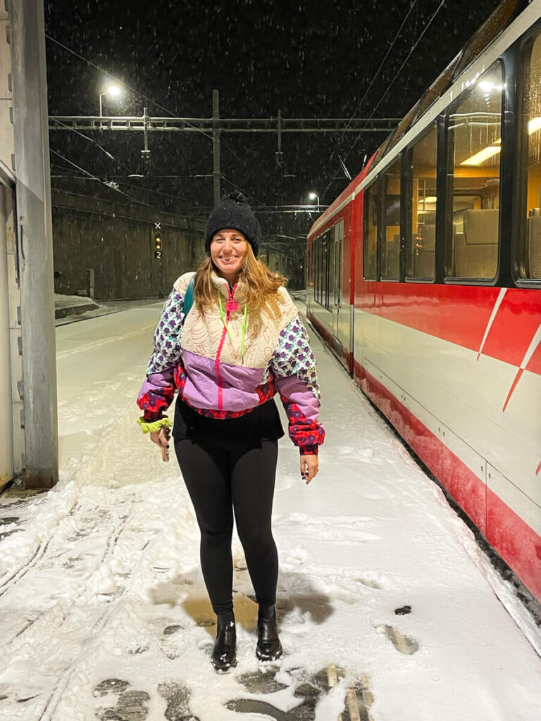 Woman in a colorful fleece jacket standing on a snowy platform at night beside a red mountain train as snow falls