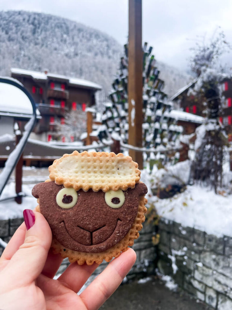 Hand holding a chocolate sheep-face cookie in front of snowy chalets in Zermatt, Switzerland