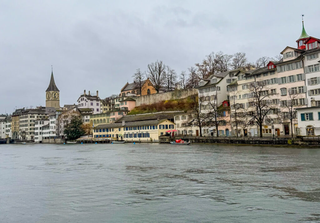 Zürich old town buildings along the Limmat River on a cloudy day, with St. Peter’s Church clock tower on the left